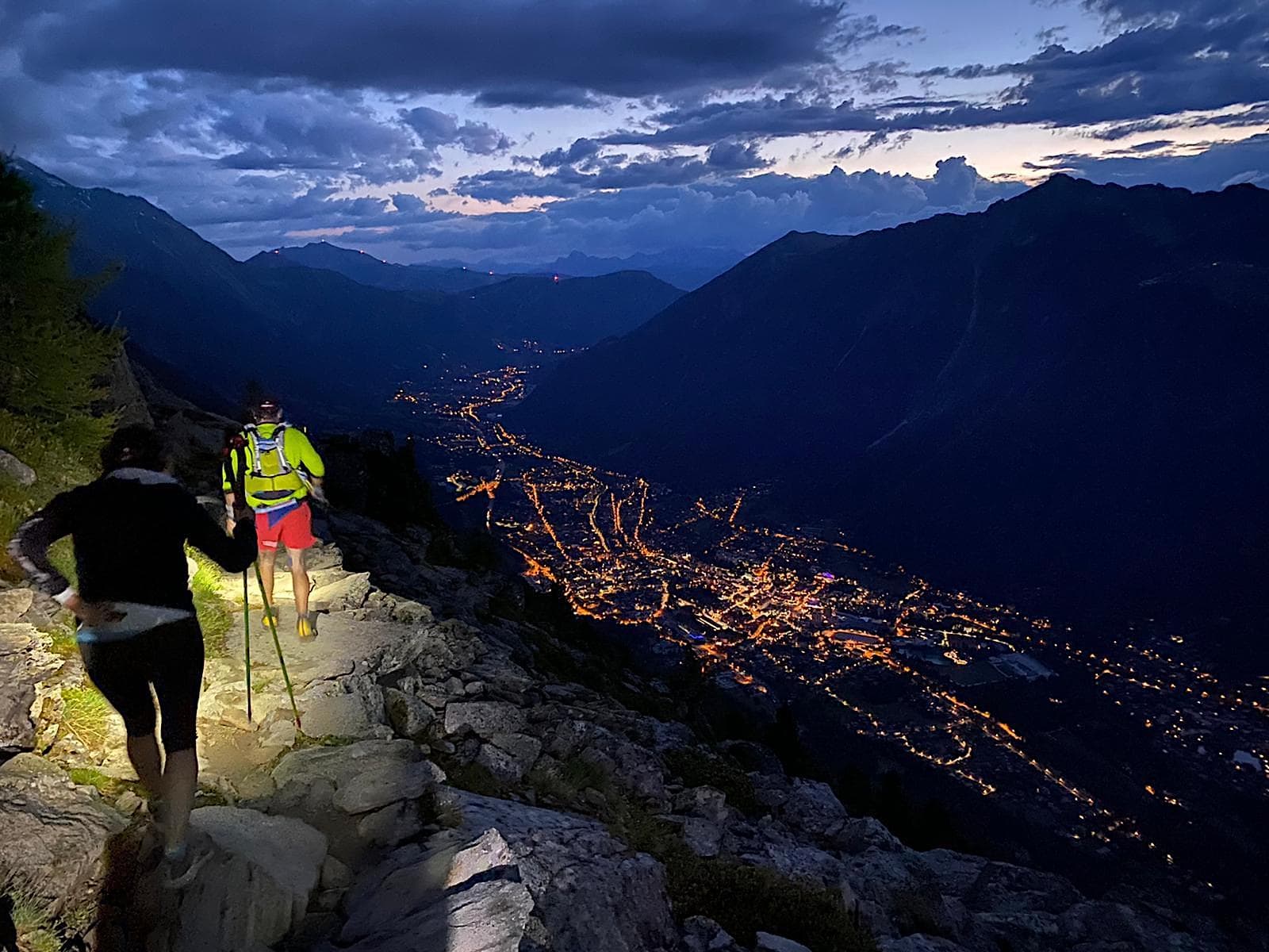 Trail running in the dark. Runners wearing headlamps on a mountain trail with Chamonix lights in the valley below. 
