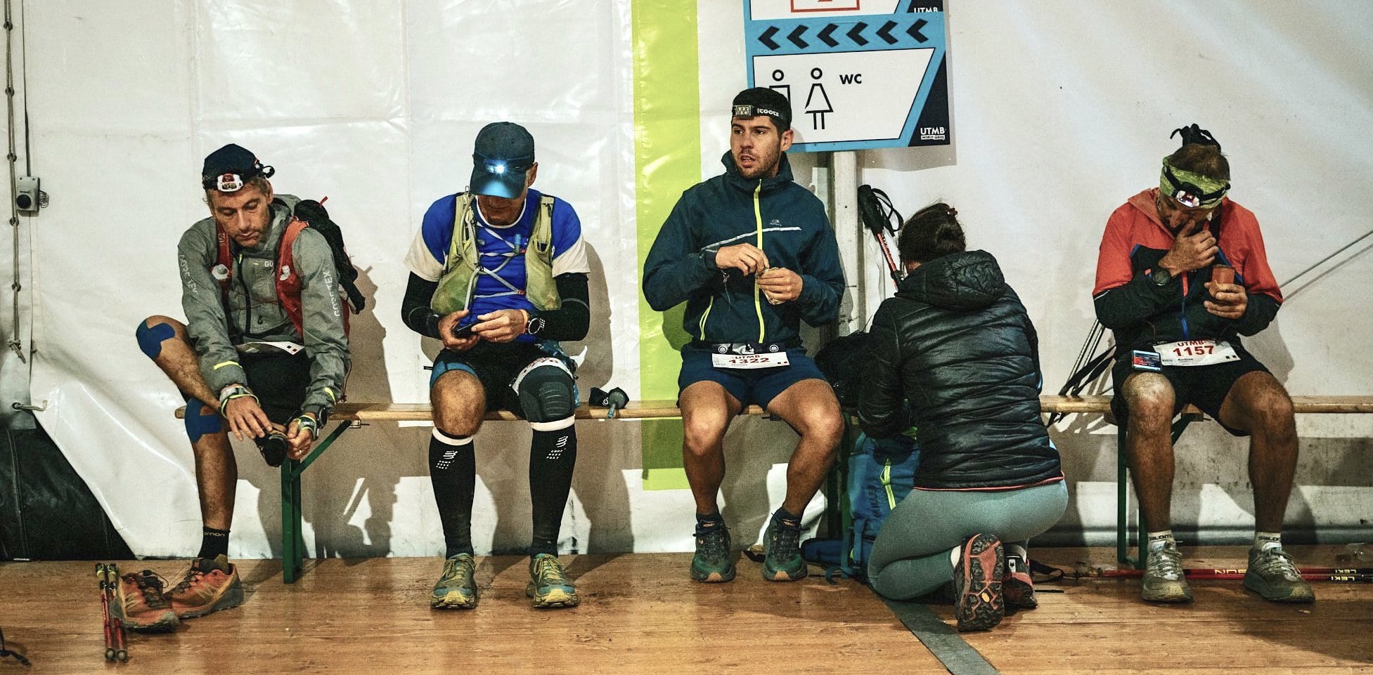 UTMB runners on a bench in an aid station, eating, sorting gear and checking feet.